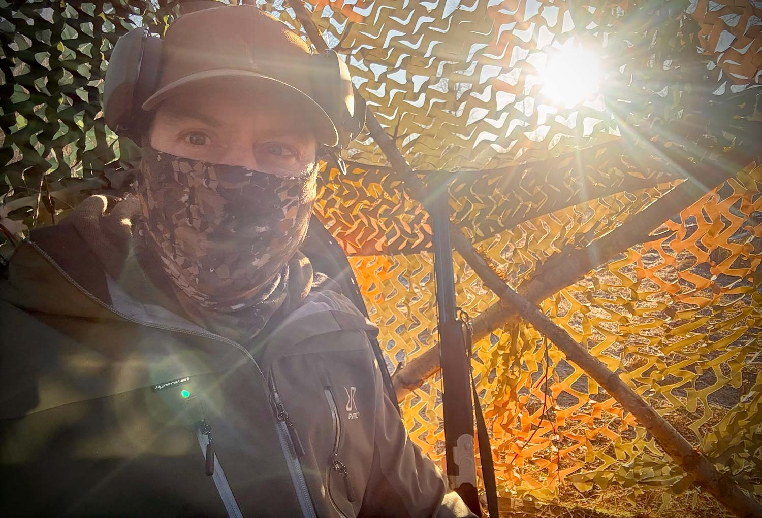 Jägare i kamouflagemönstrat gömsle under kråkjakt med ansiktsmask, RevolutionRace Silence ProShell 3L skaljacka och hörselskydd i morgonljus